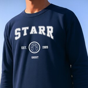 Può includere: Felpa blu navy con la scritta "STARR" a lettere bianche sul petto. Sotto, il testo "EST. 1989 GHOST" è anch'esso in bianco. La felpa ha un girocollo e maniche lunghe.