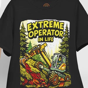 May include: Black t-shirt featuring a cartoon excavator in action, set against a forest backdrop. The text "EXTREME OPERATOR IN LIFE" is displayed in a yellow, stylized font. A white baseball cap hangs on a wooden chair.
