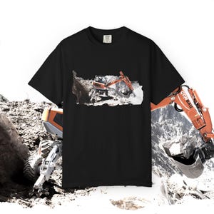 Può includere: T-shirt nero con grafica di escavatore arancione. L'escavatore è raffigurato in un cantiere. La maglietta ha un girocollo e maniche corte. L'immagine mostra anche una vista parziale di un altro escavatore.