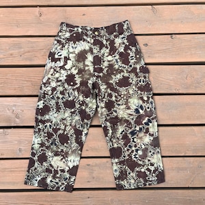 May include: Brown and cream tie-dye pants with a high waist and a button closure. The pants have a unique pattern and are made of a soft material. The pants are cropped and have pockets on the sides. The pants are laid out on a wooden surface.
