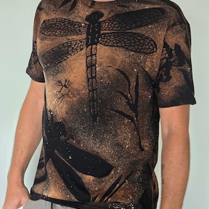 Könnte beinhalten: Schwarzes T-Shirt mit einem großen Libellen-Design. Die Libelle ist schwarz mit detaillierten Flügelmustern, vor einem braunen und beigen Hintergrund. Das T-Shirt hat ein verwaschenes, gesprenkeltes Aussehen.