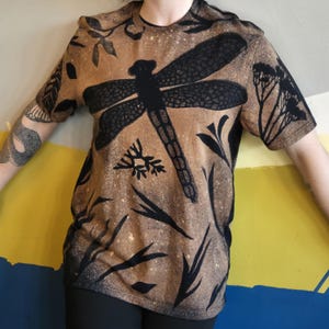 Puede incluir: Camiseta de manga corta con un diseño de libélula marrón y negro. La camiseta presenta una gran libélula negra en el centro, rodeada de patrones de plantas y hojas negras. La camiseta tiene un aspecto descolorido y vintage.