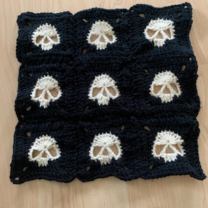 May include: A black and cream-colored crocheted blanket featuring a repeating skull pattern. The skulls are cream-colored and are set against a black background. The blanket is square-shaped and appears to be handmade.