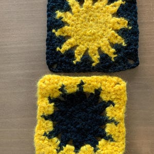 Könnte beinhalten: Zwei handgefertigte Häkelquadrate. Ein Quadrat zeigt ein gelbes Sonnendesign auf marineblauem Hintergrund. Das andere Quadrat hat ein marineblaues Sonnendesign auf gelbem Hintergrund. Die Quadrate sind aus weichem Garn gefertigt.
