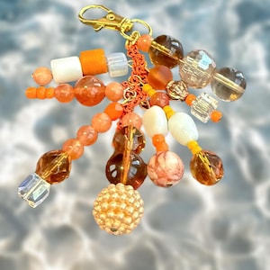 May include: A decorative charm featuring a gold-tone lobster clasp attached to a central orange braided cord. Multiple strands of beads in shades of orange, amber, white, and clear glass hang from the cord, including faceted, round, and textured beads.