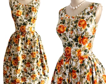 Pode incluir: Um vestido sem mangas com base branca e estampa floral de rosas laranjas e amarelas, decote redondo, corpete ajustado com pregas subtis e uma saia ampla e plissada, ajustada na cintura com um cinto fino do mesmo tecido.