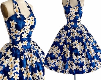 Pode incluir: Um vestido sem mangas com decote halter sobre um fundo azul profundo com um padrão repetitivo de flores de plumeria brancas e amarelas. O vestido tem um corpete justo com decote em coração e uma saia plissada ampla até aos joelhos.