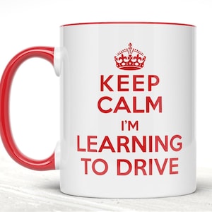 May include: A white ceramic mug with a red handle and interior rim. The front features a red graphic of a crown above the text KEEP CALM I'M LEARNING TO DRIVE in red capital letters. The design is centred on the smooth, cylindrical surface.