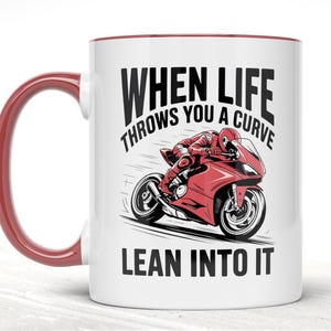 Könnte beinhalten: Weißer Keramikbecher mit rotem Henkel und Rand. Der Becher zeigt eine Grafik eines roten Motorrads und Fahrers mit dem Text "WHEN LIFE THROWS YOU A CURVE LEAN INTO IT" in Schwarz.