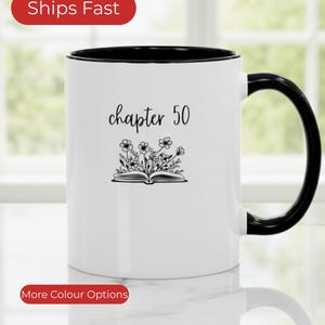 Peut inclure: Mug en céramique blanche avec intérieur et anse noirs. Le mug porte l'inscription "chapter 50" au-dessus d'une illustration de livre ouvert avec des accents floraux. Le mug est présenté sur une surface réfléchissante.