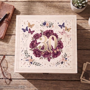 May include: A square wooden box featuring a printed design of the number 30 surrounded by a wreath of dark purple flowers and various butterflies in shades of pink, blue, and gold, enclosed within a decorative border on a light wood surface.