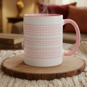 Peut inclure: Une tasse en céramique blanche avec un intérieur et une anse roses, ornée d'un motif rose répétitif. De la vapeur s'élève de la tasse, suggérant une boisson chaude. La tasse est posée sur un dessous de verre en bois, avec un arrière-plan chaleureux.