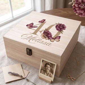 Può includere: Una scatola portaricordi quadrata in legno con chiusura in metallo, decorata con il numero 40, il nome Melissa, fiori viola e farfalle. Una piccola fotografia quadrata di una persona è esposta su un supporto in legno davanti alla scatola.