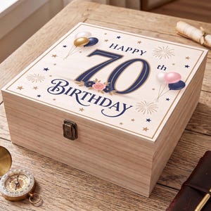 Op de afbeelding: Een vierkante houten aandenkendoos met een metalen sluiting. Het deksel bevat de tekst "HAPPY 70th BIRTHDAY" in donkerblauw, omringd door illustraties van gouden en blauwe sterren, vuurwerk en trossen roze en blauwe ballonnen en bloemen.