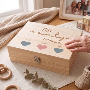 May include: A wooden keepsake box with the words "This aunty belongs to" engraved on the lid. The lid features three heart shapes in blue and pink, with names below each heart. A hand with a gold ring rests on the box.