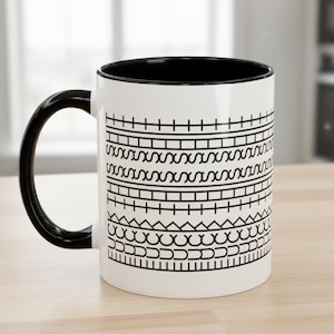 Puede incluir: Taza de cerámica blanca con interior y asa negros, con patrones geométricos negros en el exterior. Los patrones incluyen líneas horizontales, zigzags y formas curvas. La taza está sobre una superficie de madera clara.