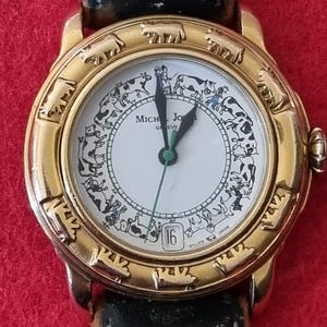 Peut inclure: Montre-bracelet dorée avec un cadran blanc orné d'un motif de vaches. Le bracelet en cuir noir est orné de broderies florales. Le cadran affiche la date et la marque "Michel Jordi Geneve".