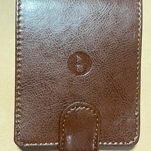 Peut inclure: Un portefeuille en cuir marron avec une surface texturée et une bordure cousue. Le portefeuille a une fermeture à rabat avec une petite languette arrondie et un emblème circulaire sur le devant. La couture est de couleur marron clair.