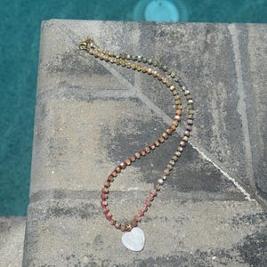 May include: A beaded necklace with a heart pendant. The necklace features small, colorful beads in shades of brown, pink, and blue. The heart pendant is white. The necklace is lying on a gray surface, with a blue pool in the background.