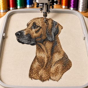 May include: An embroidery hoop holding a piece of light beige fabric with a detailed machine-stitched portrait of a brown dog with black muzzle and ear markings. The dog is shown in profile facing left, with visible needlework texture and shading.