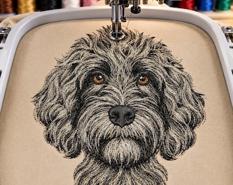 May include: An embroidery machine hoop containing beige fabric with a detailed machine-embroidered portrait of a shaggy-haired dog. The dog features textured grey and black fur, a black nose, and amber-colored eyes, positioned under the needle.