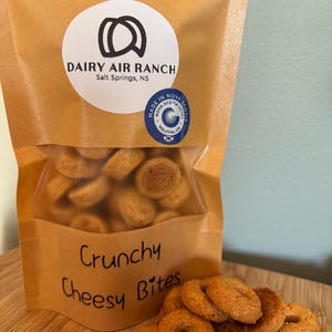 May include: A brown paper bag of "Crunchy Cheesy Bites" snacks from Dairy Air Ranch. The bag has a clear window revealing the snacks. The snacks are small, ring-shaped, and golden brown. The Dairy Air Ranch logo and "Made in Nova Scotia" seal are visible.