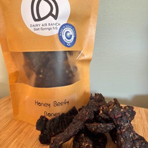 May include: A brown paper bag with a clear window filled with dark jerky. The bag features a white circular label with the text "Dairy Air Ranch Salt Springs NS" and a blue sticker that says "Made in Nova Scotia". The words "Honey Beefy Bones" are written on the bag.