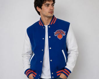 Könnte beinhalten: Eine blau-weiße New York Knicks Collegejacke. Die Jacke hat einen blauen Körper mit weißen Ärmeln, orange-blau gestreiften Besätzen und ein Knicks-Logo auf der Brust. Sie hat eine Knopfleiste.