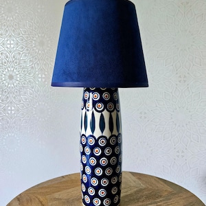 Op de afbeelding: Een tafellamp met een cilindrische keramische voet versierd met een herhalend patroon van donkerblauwe en witte cirkels met oranje centra. De lamp is voorzien van een effen donkerblauwe stoffen lampenkap met een taps toelopende, conische vorm.