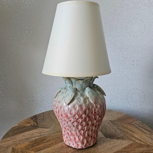 Może przedstawiać: Ceramiczna lampa stołowa w kształcie truskawki. Czerwona podstawa ma białe plamki i zielone liście. Biały abażur jest umieszczony na górze. Lampa stoi na drewnianym stole. Wysokość około 30 cm.