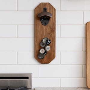May include: A wooden bottle opener with a dark brown finish, mounted on a white tiled wall. The opener has a bronze-colored mechanism and is holding several bottle caps. The design is rectangular with a unique shape.