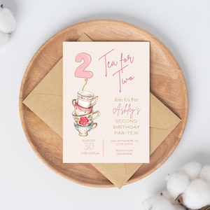 Puede incluir: Una tarjeta de invitación rosa claro con el texto "Tea for Two" y "Ashley's Second Birthday Par-Tea!". Presenta una pila de tazas de té y un globo rosa con el número 2. La invitación está sobre un plato de madera con un sobre marrón.