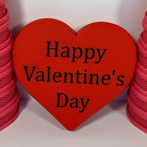 May include: A Valentine's Day themed image featuring two stacks of pink, textured, heart-shaped boxes flanking a red heart. The red heart in the center has the words "Happy Valentine's Day" printed in black.