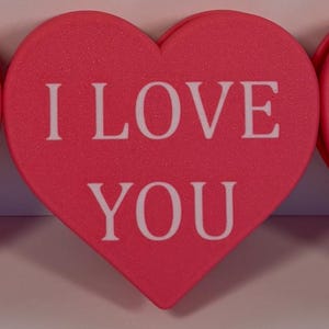 May include: Four pink heart-shaped decorations with white text: "BE MY VALENTINE", "FOR THE SWEETEST GIRL", "I LOVE YOU", and "HAPPY VALENTINE'S DAY". A red can is on the right. Valentine's Day decor.