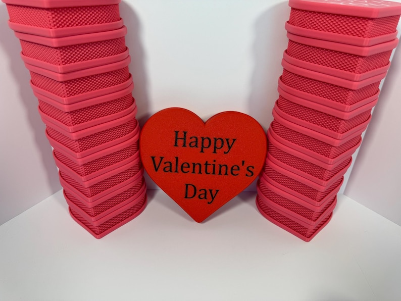 May include: A Valentine's Day themed image featuring two stacks of pink, textured, heart-shaped boxes flanking a red heart. The red heart in the center has the words "Happy Valentine's Day" printed in black.