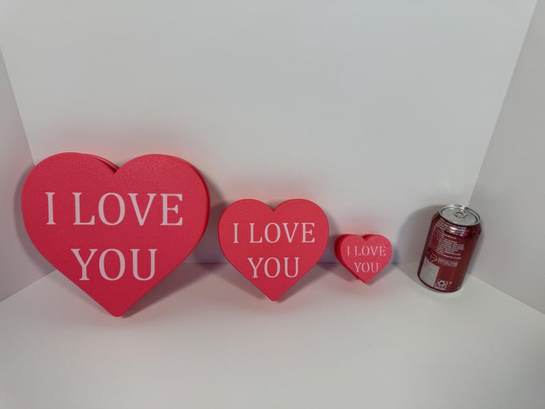 May include: Three pink heart-shaped decorations with the text "I LOVE YOU" in white. A red soda can is next to the hearts. The hearts are different sizes, with the largest on the left.