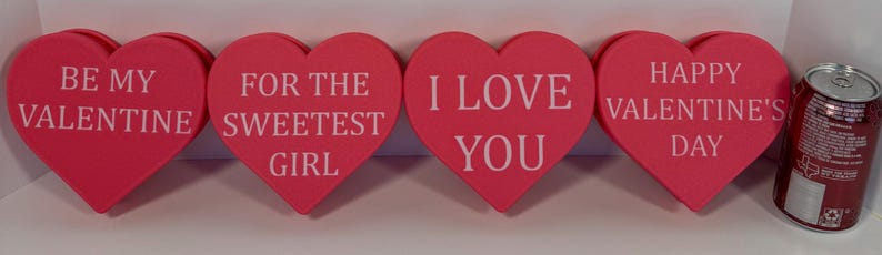 May include: Four pink heart-shaped decorations with white text: "BE MY VALENTINE", "FOR THE SWEETEST GIRL", "I LOVE YOU", and "HAPPY VALENTINE'S DAY". A red can is on the right. Valentine's Day decor.