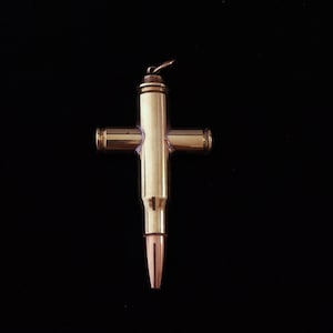 May include: A brass-colored cross pendant made from repurposed bullet casings. The cross features a central vertical casing with three horizontal casings attached, forming the cross shape. The bullet tips are copper-colored.