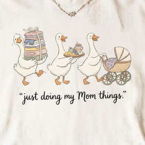 Peut inclure: Un t-shirt à manches courtes blanc cassé avec une illustration de trois oies blanches. Une oie porte une pile de vêtements pliés, une autre tient un plateau de nourriture et la troisième pousse un landau. Le texte indique "just doing my Mom things."