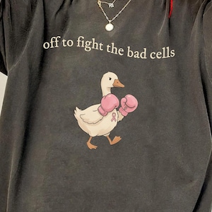 May include: A charcoal gray short-sleeve t-shirt featuring a graphic of a white duck wearing pink boxing gloves. The text "off to fight the bad cells" is printed in a light beige font above the duck illustration, which includes a small pink ribbon.
