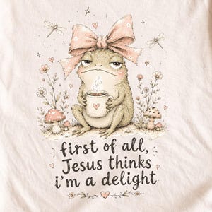May include: A light beige t-shirt featuring a graphic of a frog wearing a large pink bow, holding a steaming mug with a heart icon. The design includes mushrooms, dragonflies, and the text: first of all, Jesus thinks i'm a delight.