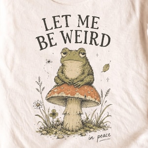 May include: A light beige t-shirt featuring a graphic illustration of a green frog sitting on a large orange and white mushroom. The text "LET ME BE WEIRD" is printed in black above the frog, and the words "in peace" are printed below the mushroom.
