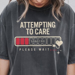 May include: A charcoal gray short-sleeve t-shirt featuring the text ATTEMPTING TO CARE in beige, a loading bar graphic with three red segments, the word LOADING, a pixelated heart icon, and the text PLEASE WAIT with three red dots.