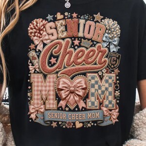 Op de afbeelding: Een zwart t-shirt met een grafisch ontwerp met de tekst SENIOR CHEER MOM. Het ontwerp bevat een groot centraal hart met een strik, pompons, sterren en ruitpatronen in tinten roze, beige en blauw, met een banner aan de onderkant.