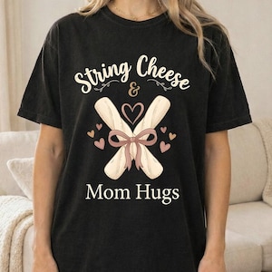 May include: Black t-shirt with the text "String Cheese & Mom Hugs". The design shows two string cheese sticks tied with a bow and hearts. The shirt is a relaxed fit. The text and design are in a playful style.
