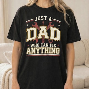 Puede incluir: Camiseta negra con el texto "JUST A DAD WHO CAN FIX ANYTHING" en un diseño desgastado. El gráfico incluye llaves inglesas y estrellas. La camiseta es de corte holgado.