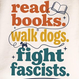 Könnte beinhalten: Ein cremefarbenes Sweatshirt mit Rundhalsausschnitt und dem Text "read books. walk dogs. fight fascists." in Orange, Gelb und Dunkelblau. Das Design enthält eine kleine Strichzeichnung eines offenen Buches und die Silhouette eines Hundes an der Leine.