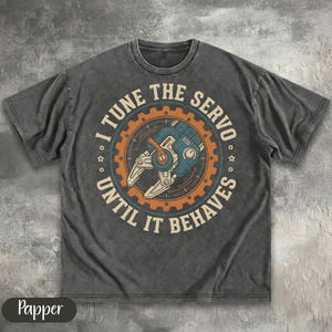 May include: A charcoal grey short-sleeve t-shirt featuring a circular graphic with a mechanical servo claw in the centre. The text "I TUNE THE SERVO UNTIL IT BEHAVES" is printed in a bold, arched font around the orange and blue gear-shaped border.