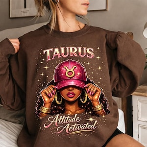 May include: A brown sweatshirt featuring a graphic of a woman wearing a pink Taurus cap. The word "TAURUS" is above the image, and "Attitude Activated" is below. The design includes gold accents and star details.