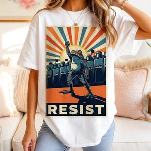 May include: White t-shirt featuring a frog illustration with a raised fist, set against a backdrop of a rising sun and riot police. The word "RESIST" is prominently displayed below the image. The shirt is a statement piece.